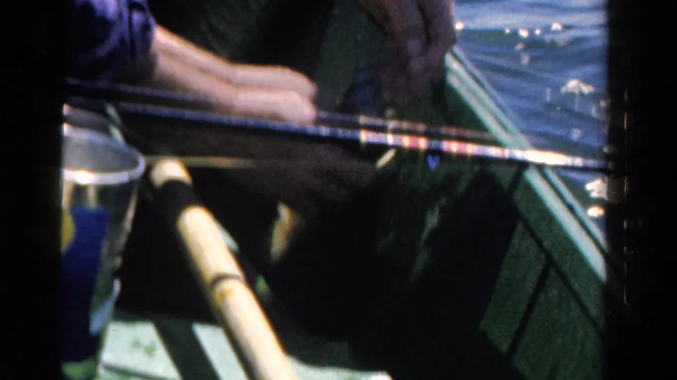 1958 Ontario 8mm frame showing a man's hands and fishing rod inside a green boat beside sunlit lake water.