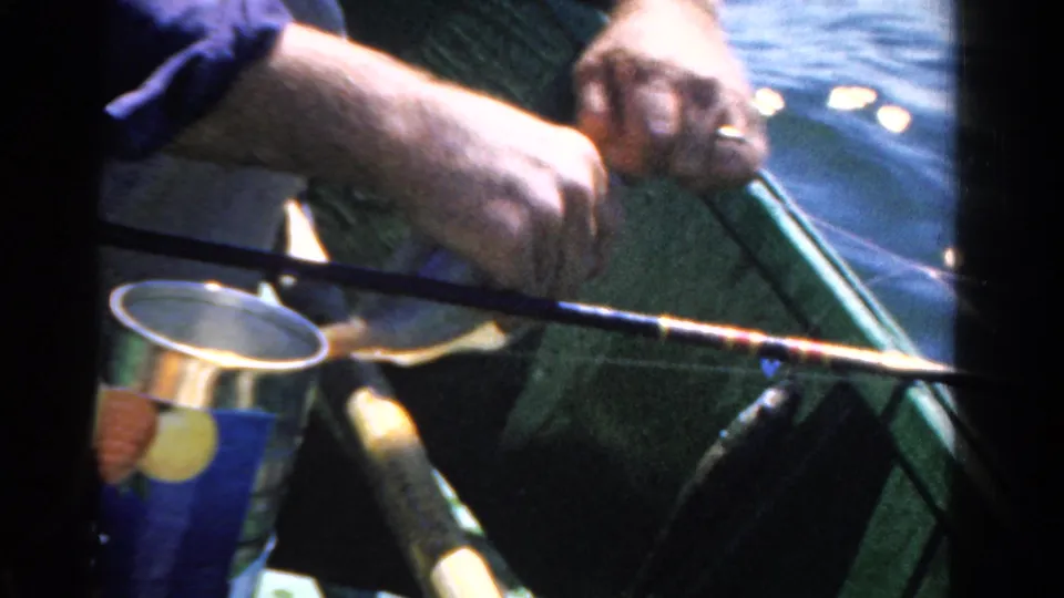 1958 Ontario home-movie frame of a man handling fishing tackle in a green boat, with calm water and a metal can visible.