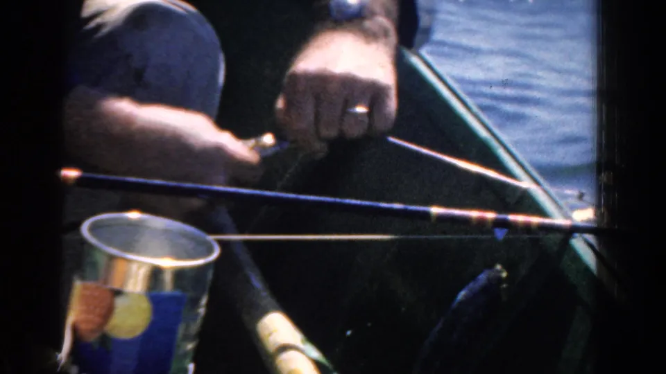 1958 Ontario home-movie frame showing a man's wedding ring as he handles fishing line beside the rod in a green boat.