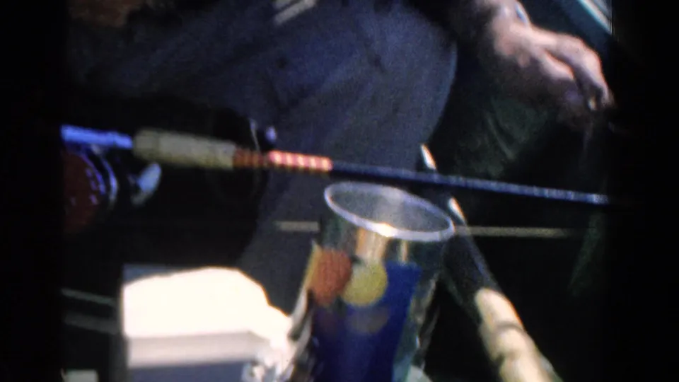 Close 8mm view of a fishing rod crossing the frame, with a bait can and part of the seated adult visible in the boat.