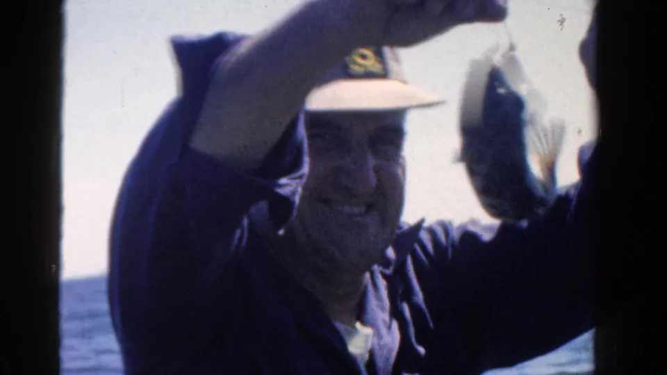 A smiling adult man in a light cap holds up his catch toward the camera in 1958 Ontario lake footage.
