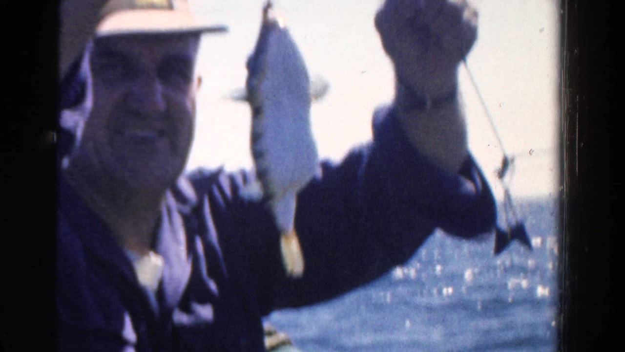 In 1958 Ontario home-movie footage, a man in a light hat smiles while displaying a fish on a line near open water.
