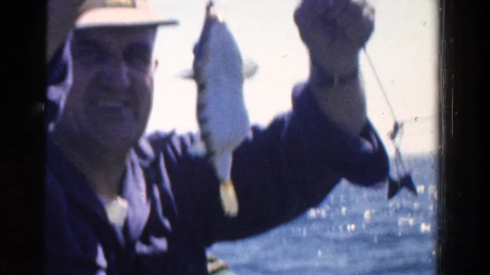In 1958 Ontario home-movie footage, a man in a light hat smiles while displaying a fish on a line near open water.