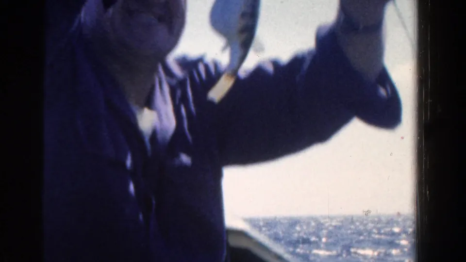 The man smiles toward the camera while holding up a small fish, with sun glare and lake water behind him.