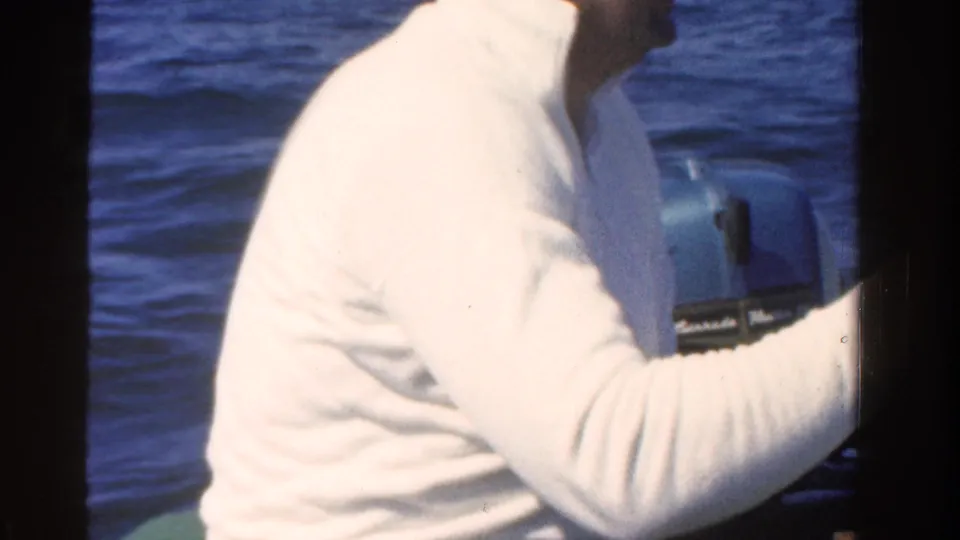 A man in a white sweater sits at the stern and handles a small outboard motor with open water behind him.