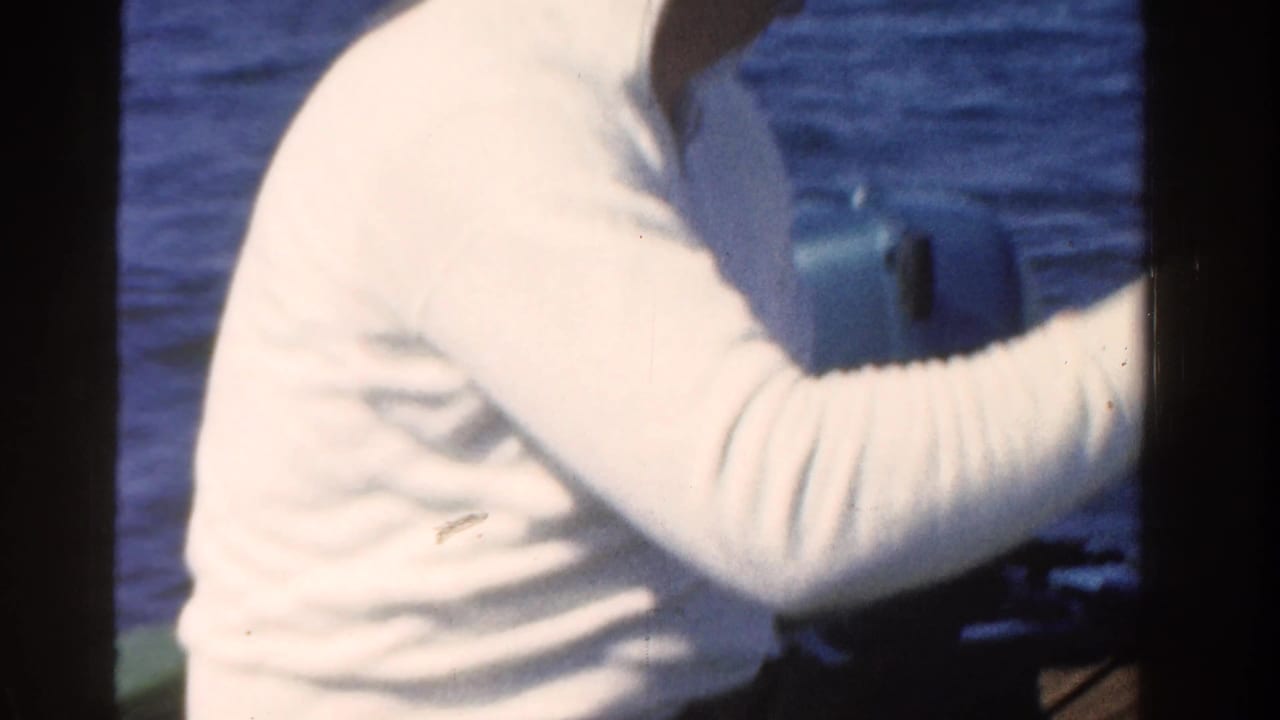 Man in a white long-sleeved shirt holds a fishing rod over blue water from a boat.