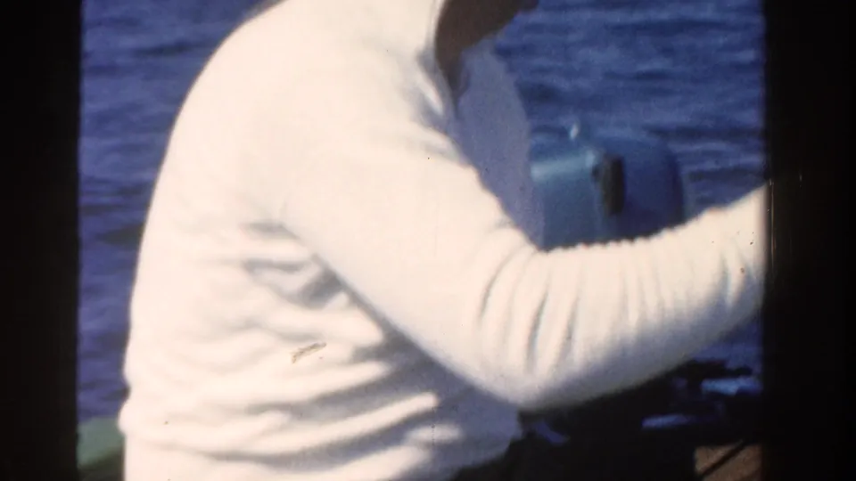 In 1958 Ontario home-movie footage, a man in a white shirt sits in a boat and works a fishing rod over open blue water.