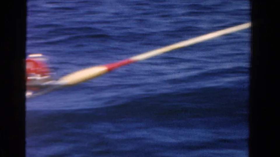A fishing rod with a red reel extends across rippled water in this 1958 Ontario 8mm fishing scene.