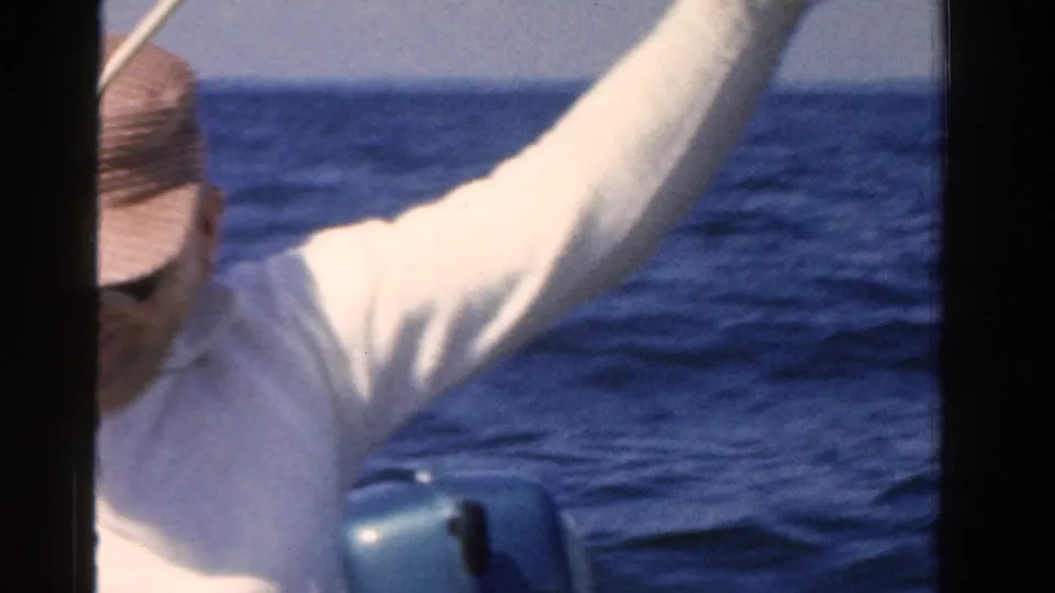 The fisherman raises his rod from a boat on Ontario water, with an outboard motor visible behind him in the 1958 home-movie frame.