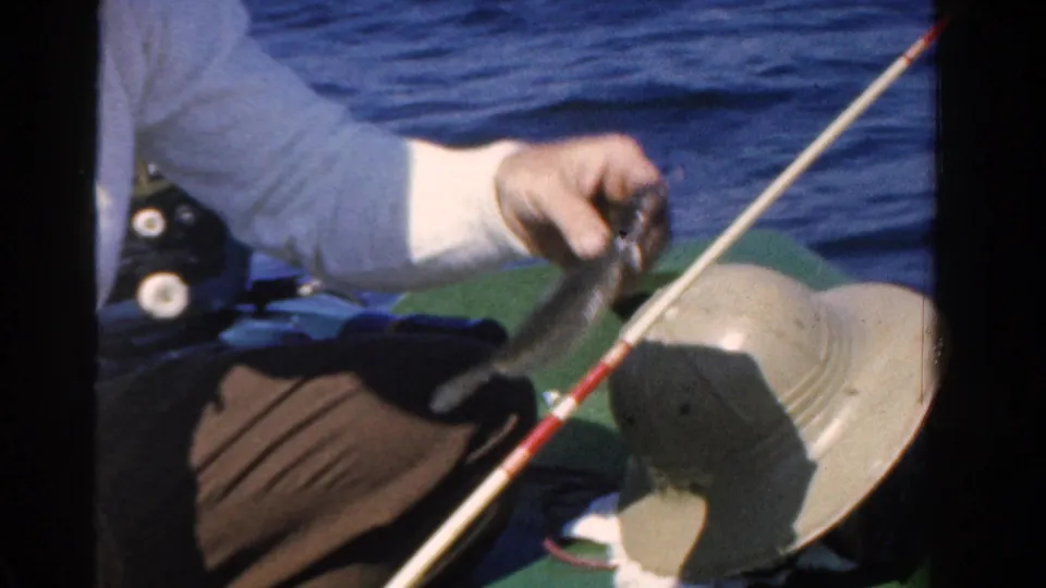 The man handles the caught fish over the green boat surface, with a fishing rod crossing the frame and blue water behind him.