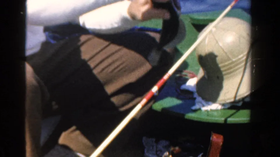 A close, slightly soft 8mm view shows the fishing rod across the boat, the man's clothing, a brimmed hat, and blue water beyond.