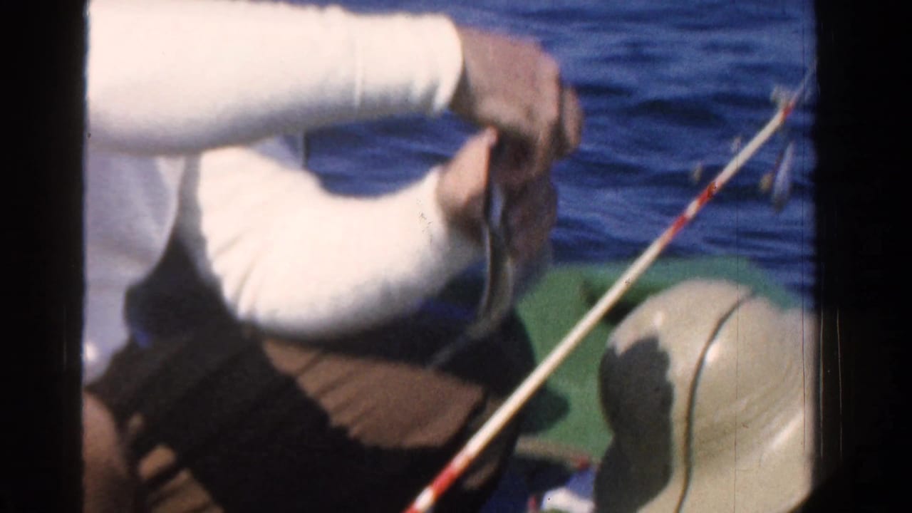 Close 1958 Ontario 8mm frame of a man handling a fish near a fishing rod on a small boat, with blue water behind him.