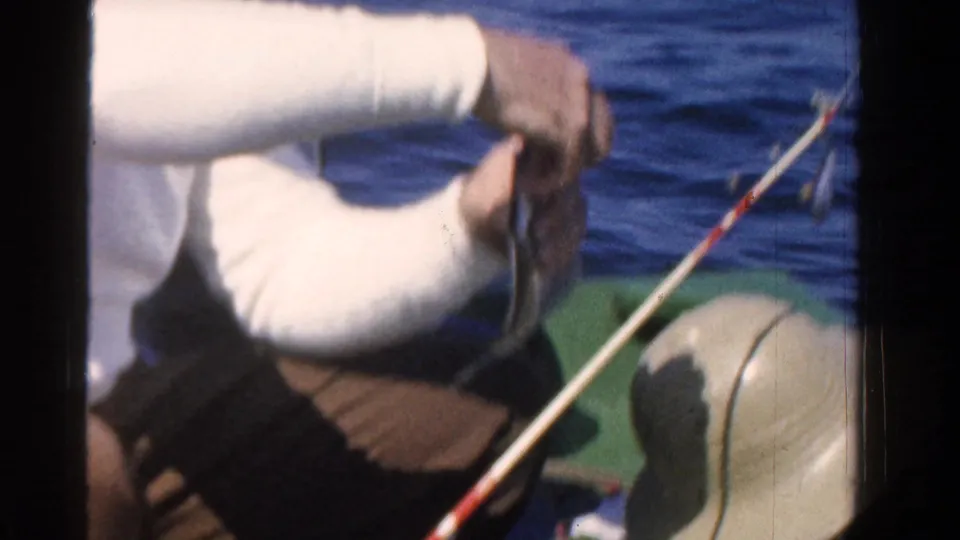 Close 1958 Ontario 8mm frame of a man handling a fish near a fishing rod on a small boat, with blue water behind him.