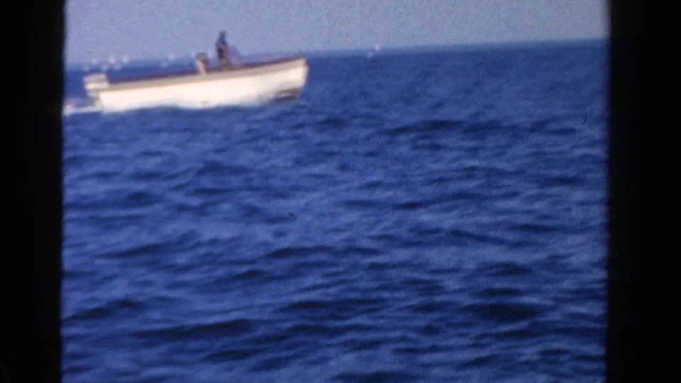 A distant small white motorboat moves across blue water, seen in soft 1958 Ontario 8mm home-movie footage.