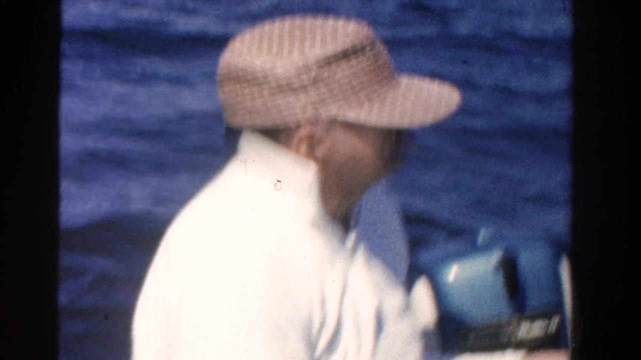 Man in a visor hat and white sweater beside a blue outboard motor on open water.