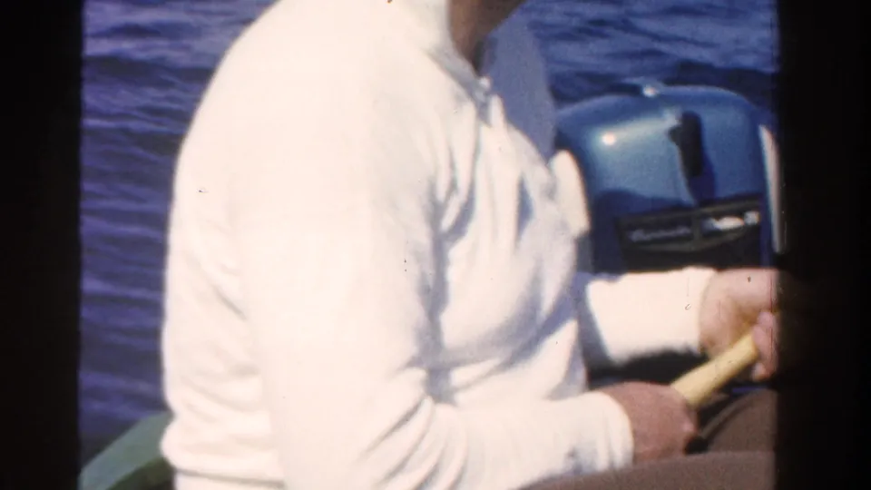 Close 1958 Ontario home-movie view of a seated man in a white sweater holding a fishing rod beside a blue outboard motor.