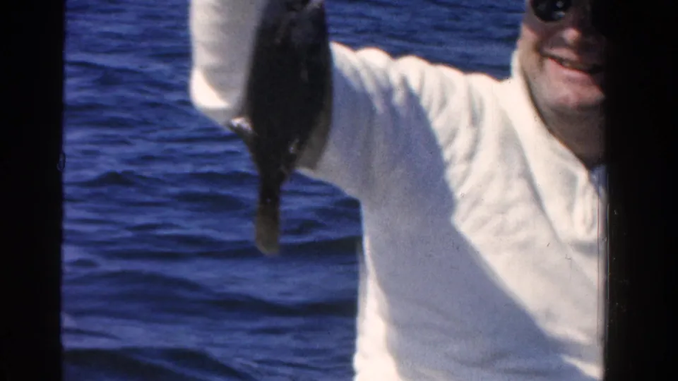 In 1958 Ontario home-movie footage, a man in sunglasses and a light cap raises a fish while seated near open water.