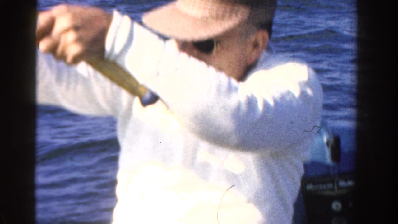 Man in a white shirt holds a fishing rod in a small boat on blue water.