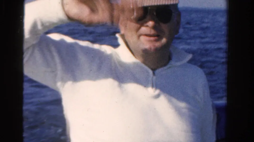 An adult man in sunglasses, straw hat, and white shirt waves from a boat in Ontario in this 1958 8mm frame.