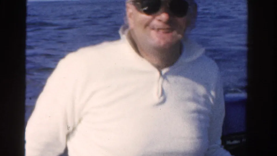 1958 Ontario 8mm frame of a smiling adult man in sunglasses and a white shirt on a boat in bright daylight.