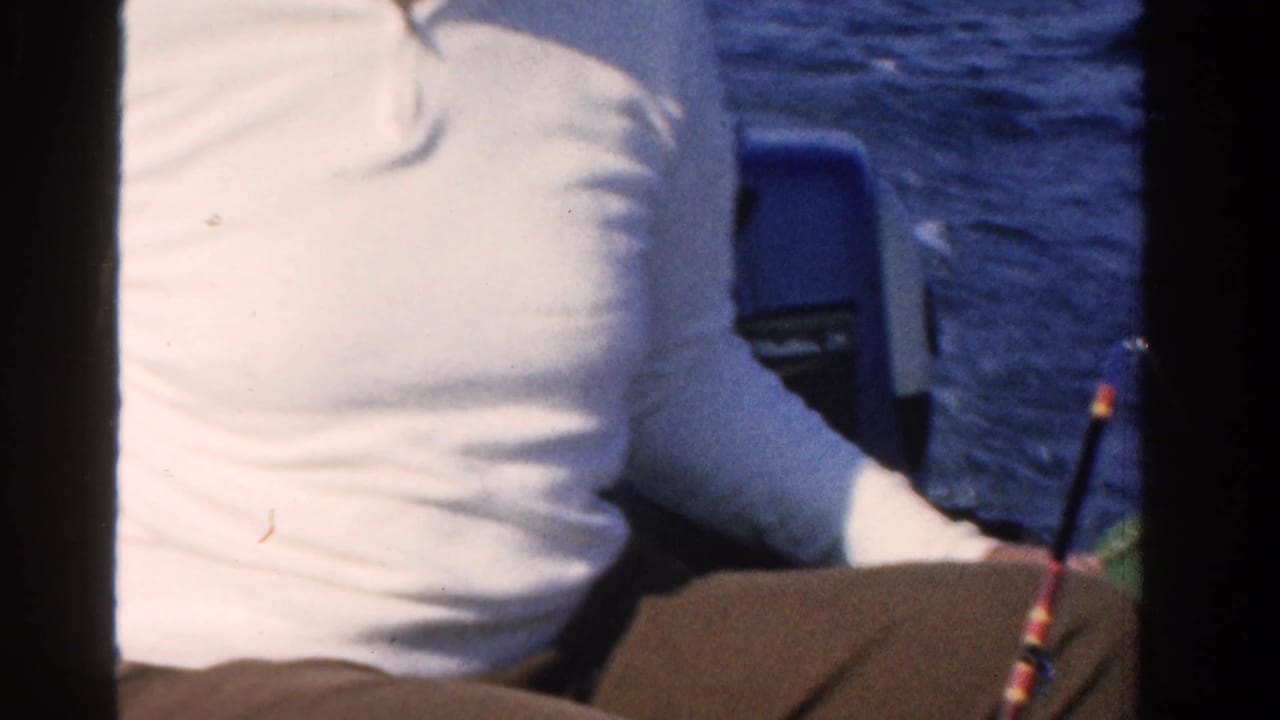 Man in a white shirt seated in a boat beside blue open water.