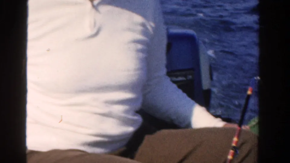 Adult man in a white shirt sits low in a boat on Ontario open water, with a fishing rod partly visible in this 1958 8mm frame.
