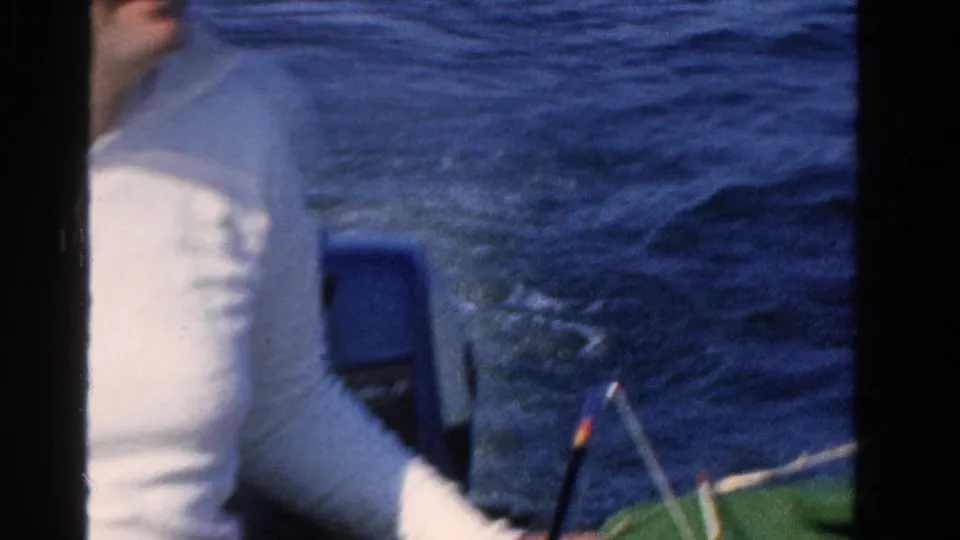 Partial 1958 Ontario boating frame showing a seated adult man in a white shirt, blue water, boat seating, and a fishing rod near the edge.