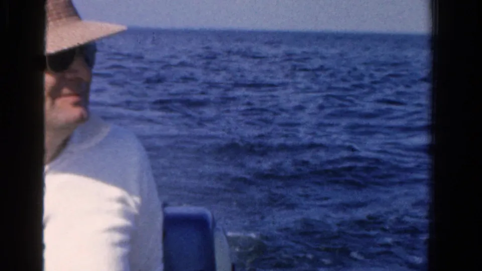 Adult man wearing a light hat and sunglasses sits in a boat against open Ontario water in a 1958 home-movie frame.