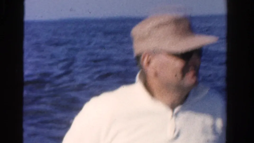 Adult man in a straw hat, sunglasses, and white shirt sits on a boat with rippled Ontario water behind him in 1958 footage.
