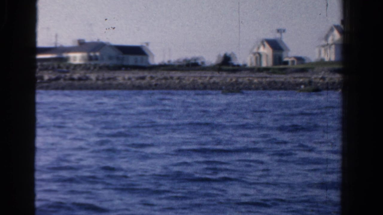 Boat view across blue water toward low white shoreline buildings under a pale sky.