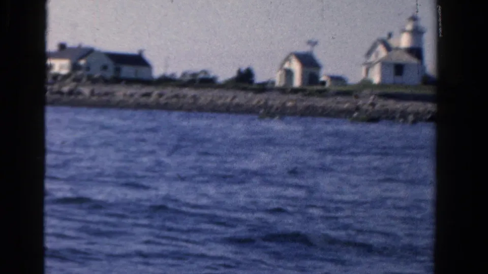 The boat passes a rocky Ontario shoreline with white houses and a small lighthouse-like waterfront structure visible in soft 8mm color.