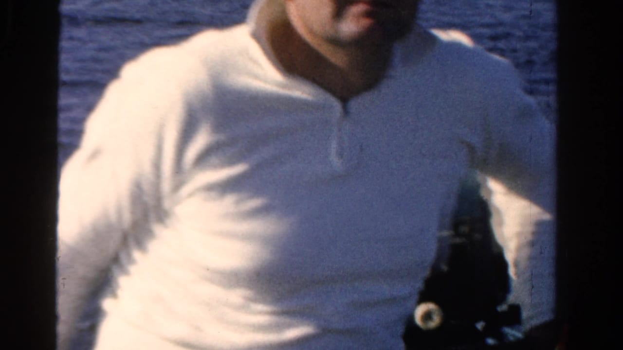 Man in a white shirt stands close to the camera on a boat with lake water behind him.