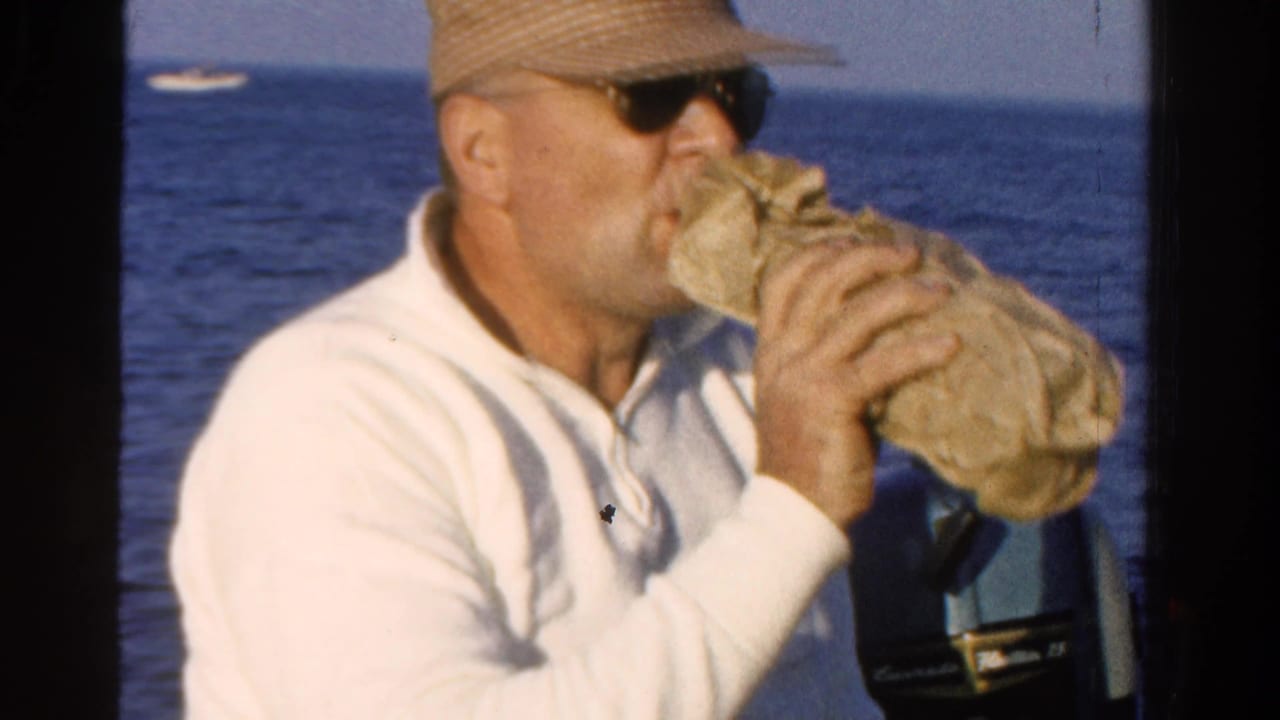 In 1958 Ontario home-movie footage, a man in a white polo, cap and sunglasses drinks from a cloth-wrapped bottle on open water.