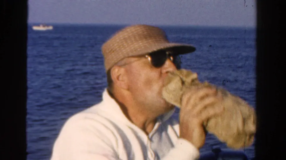 The man continues a long drink from the cloth-wrapped bottle during the 1958 Ontario boating scene, with a distant boat on the horizon.