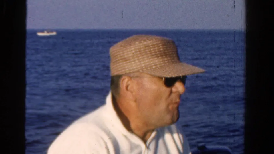 After drinking, the man lowers the bottle and looks down in the 1958 Ontario boating footage, with blue water filling the background.
