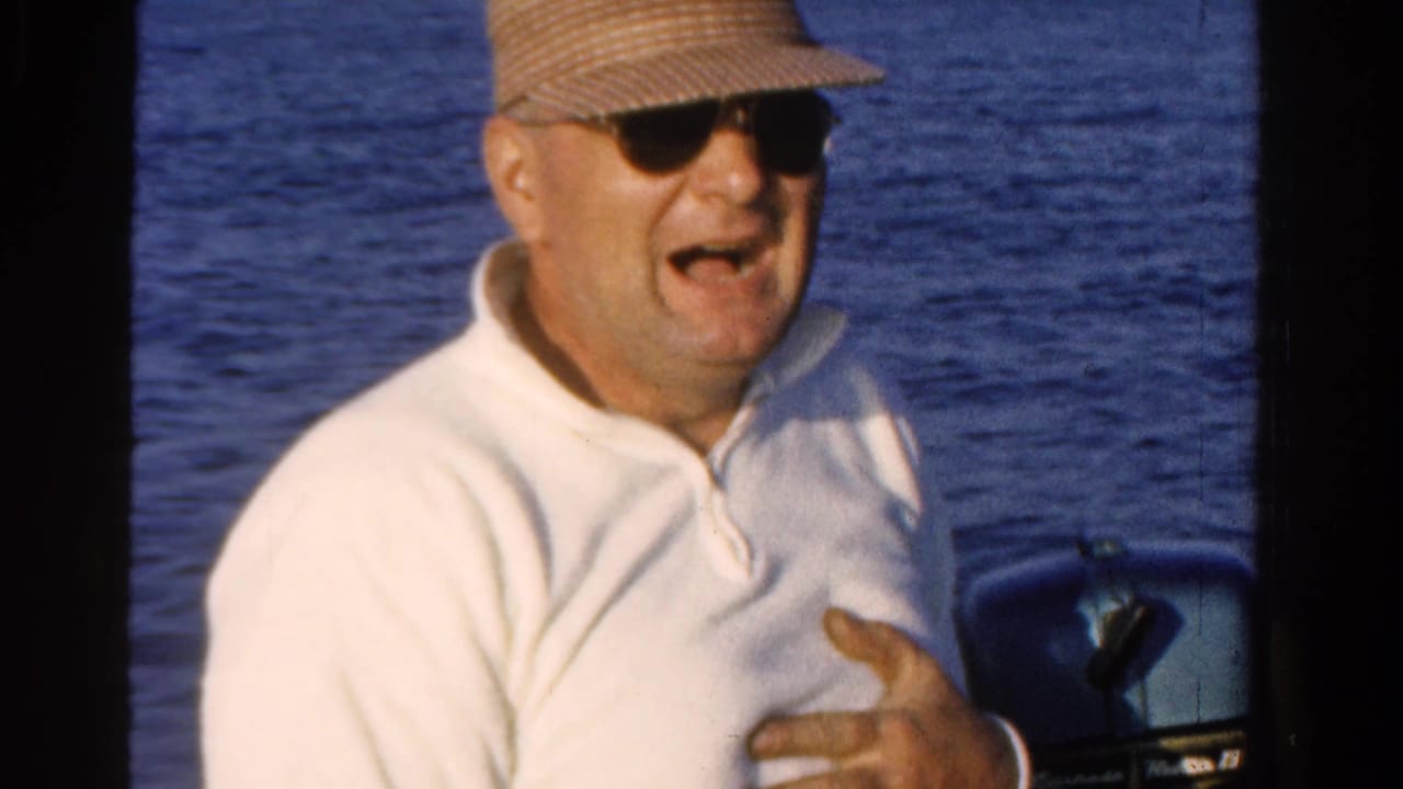 Man in sunglasses and straw cap sits on a boat with blue water behind him.