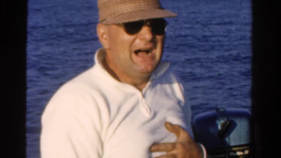 Adult man in Ontario, Canada, 1958, wearing sunglasses, straw cap, and white pullover while seated on a boat.