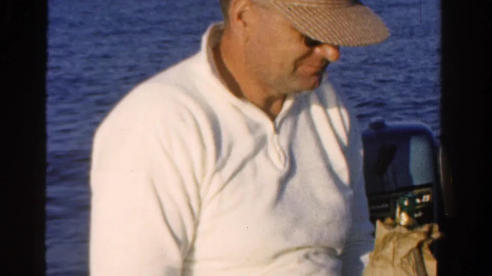Adult man on a boat in Ontario, Canada, 1958, looking down beside an outboard motor with rippling water behind him.