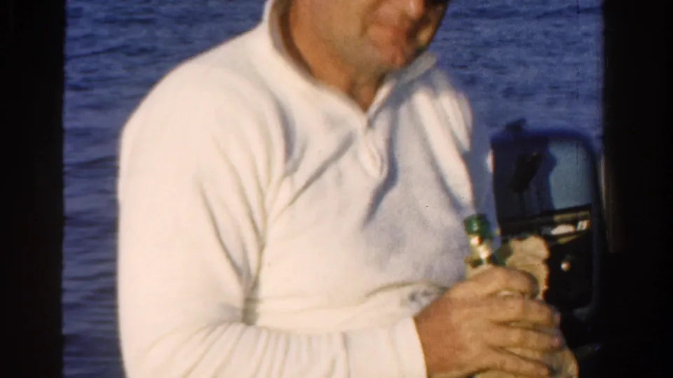 Adult man in 1958 Ontario home-movie footage holds a wrapped bottle while seated on a boat near blue water.