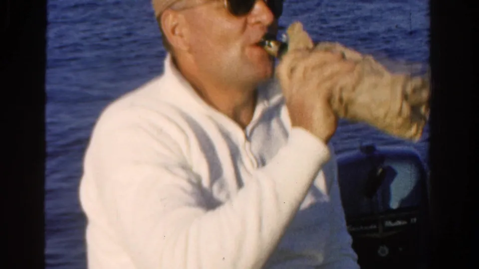 Adult man in Ontario, Canada, 1958, drinks from a wrapped bottle while seated on a boat beside an outboard motor.