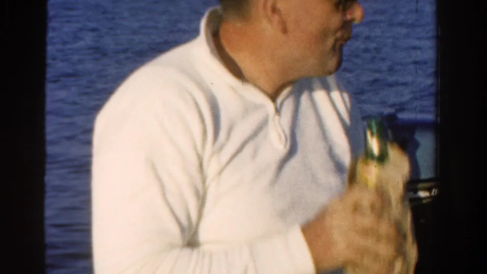 A man in a white shirt holds a bottle and turns sideways on a boat in Ontario, with open water behind him.