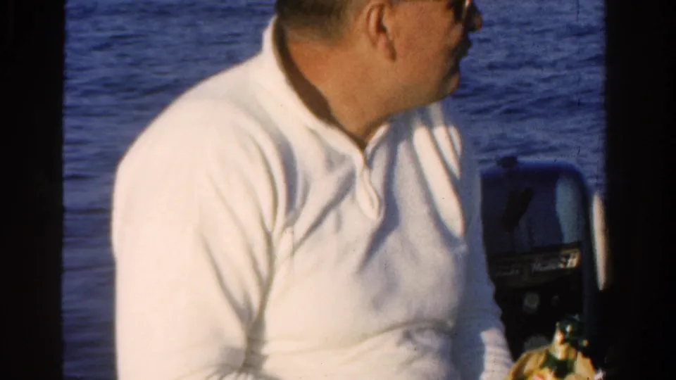 A seated man in a white pullover holds a bottle near his lap on a boat during a 1958 Ontario home-movie moment.