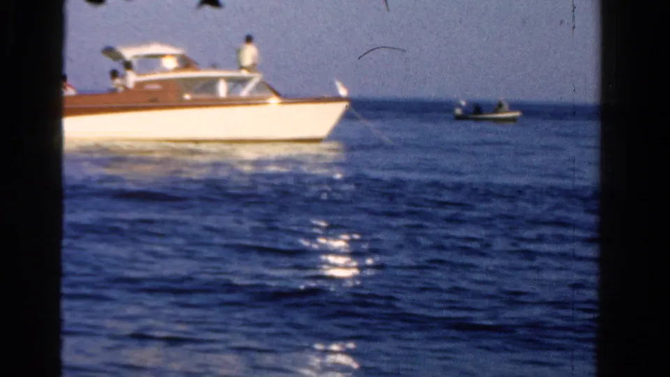 A close pass of the motorboat shows sun glare on the water and a small distant craft near the horizon.