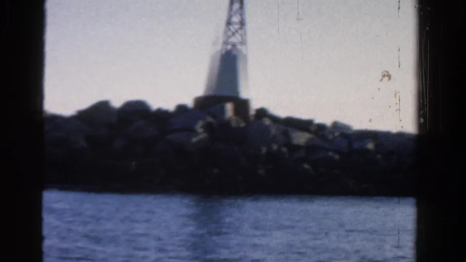 1958 Ontario home-movie frame showing a white lighthouse on rocks beyond calm blue water, with dark film borders visible.