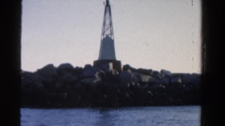 The lighthouse remains in view on the rocky shoreline, filmed across water in soft 8mm color with heavy frame edges.