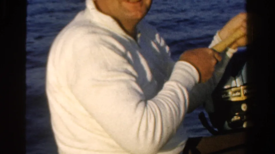 An adult man in a white shirt handles a wooden oar or boat control beside an outboard motor on the water in Ontario footage.