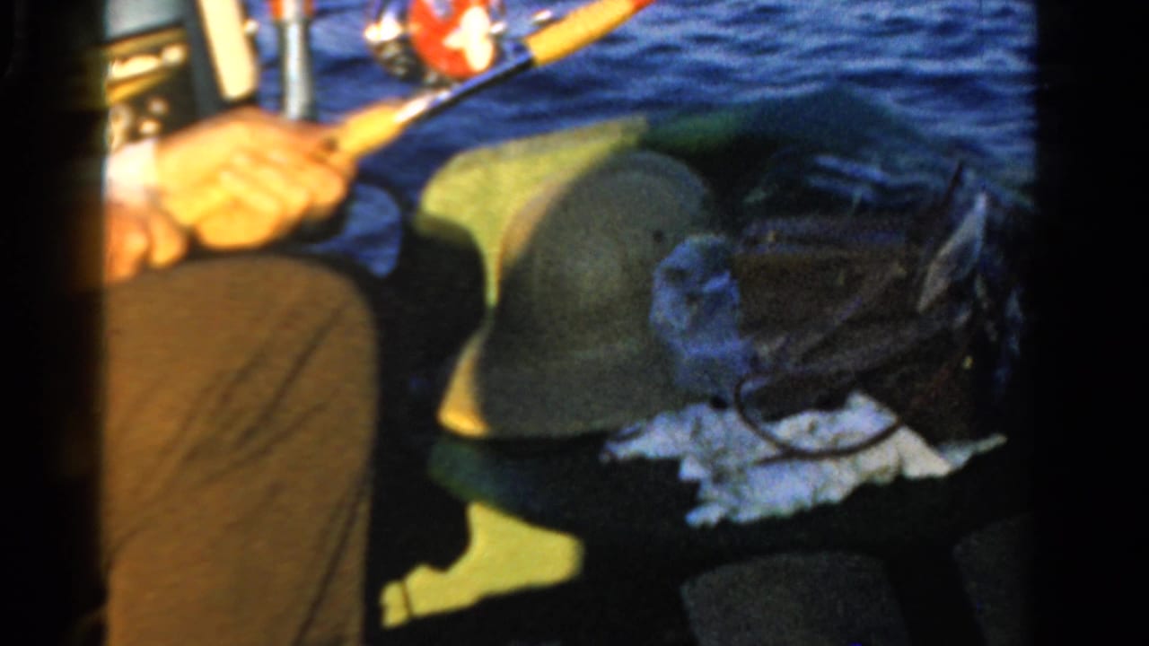 Seated fisherman holds a rod beside a green hat, bag, and rippling blue water.