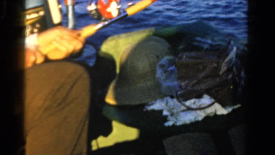 1958 Ontario home-movie frame showing a seated adult fisherman with rod, green hat, bag, and open water behind him.