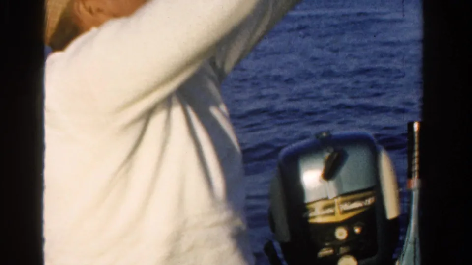 1958 Ontario home-movie frame showing the fisherman's torso near an outboard motor, rod, and open water.