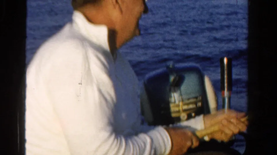 1958 Ontario frame showing an adult fisherman seated by a vintage outboard motor with blue water in the background.